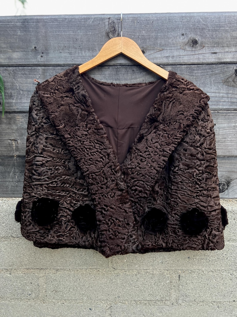 Pu&ograve; includere: Giacca marrone in stile vintage con un motivo riccio e strutturato. La giacca ha un collo a scialle e tre decorazioni scure e rotonde sul davanti. &Egrave; appesa a una gruccia di legno su uno sfondo di legno invecchiato.