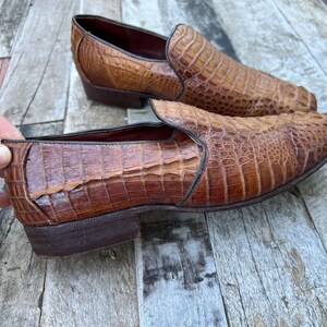 May include: A pair of brown leather slip-on shoes with a crocodile skin texture. The shoes have a low heel and a rounded toe. The leather has a rich, textured appearance, and the shoes are set against a wooden surface.