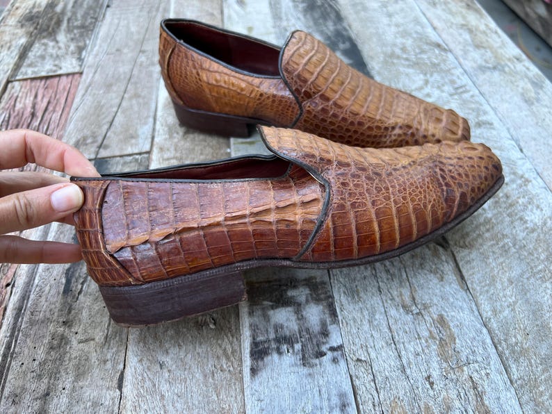 May include: A pair of brown leather slip-on shoes with a crocodile skin texture. The shoes have a low heel and a rounded toe. The leather has a rich, textured appearance, and the shoes are set against a wooden surface.