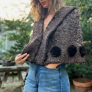 Pu&ograve; includere: Bolero marrone strutturato con collo a scialle e tre applicazioni floreali nere. La giacca &egrave; indossata sopra jeans blu. Il bolero &egrave; un modello corto e il tessuto ha una trama riccia unica.