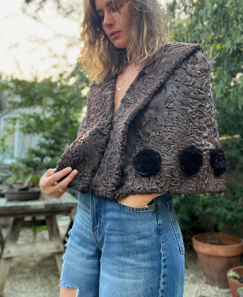 Pu&ograve; includere: Bolero marrone strutturato con collo a scialle e tre applicazioni floreali nere. La giacca &egrave; indossata sopra jeans blu. Il bolero &egrave; un modello corto e il tessuto ha una trama riccia unica.