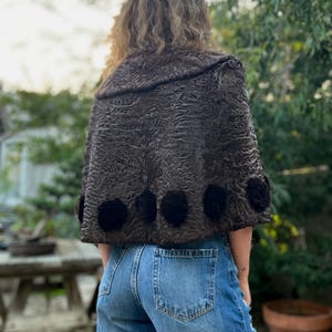 Pu&ograve; includere: Un mantello marrone scuro strutturato con colletto arrotondato e dettagli floreali neri. Il mantello &egrave; indossato sopra jeans blu. L'immagine &egrave; scattata all'aperto con un tavolo di legno e vegetazione sullo sfondo.