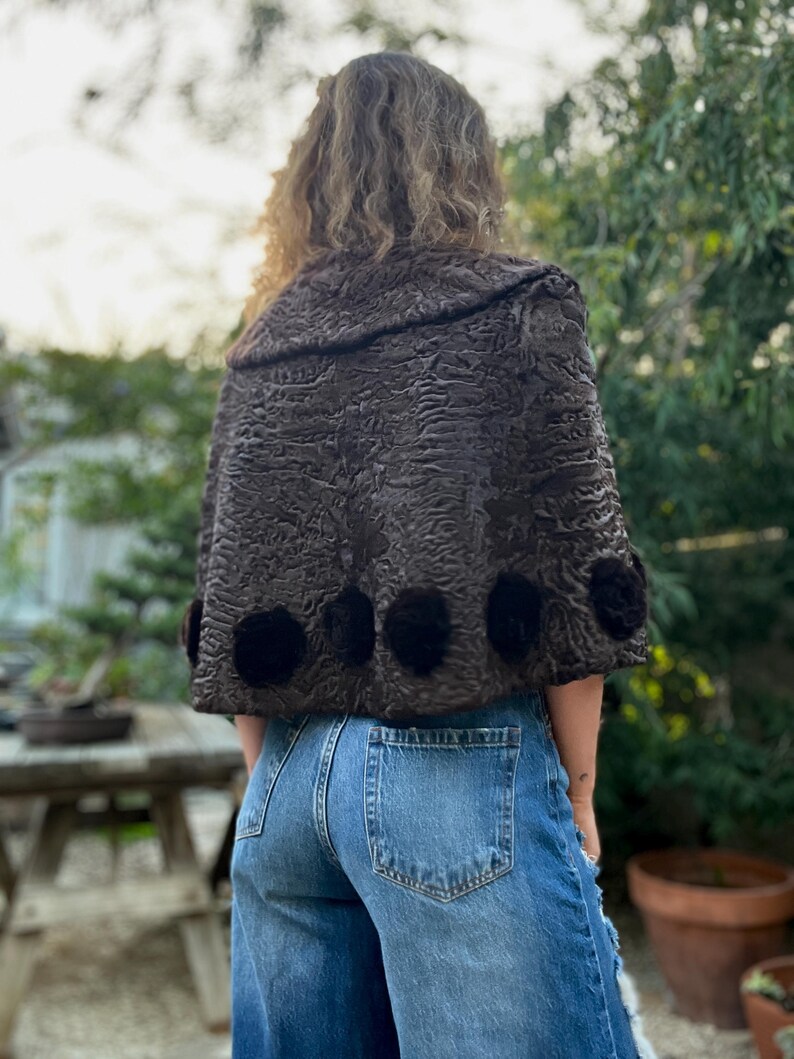 Pu&ograve; includere: Un mantello marrone scuro strutturato con colletto arrotondato e dettagli floreali neri. Il mantello &egrave; indossato sopra jeans blu. L'immagine &egrave; scattata all'aperto con un tavolo di legno e vegetazione sullo sfondo.