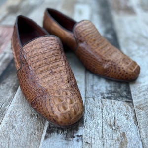 May include: A pair of brown leather loafers with a crocodile skin texture. The shoes have a classic slip-on design with a rounded toe and a dark brown trim. The loafers are displayed on a wooden surface.