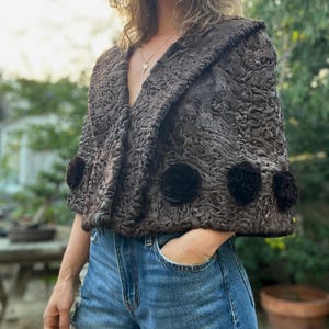 Pu&ograve; includere: Un mantello marrone strutturato con collo a scialle e tre accenti floreali scuri. Il mantello &egrave; indossato sopra pantaloncini di jeans blu. L'immagine &egrave; scattata all'aperto con luce naturale.