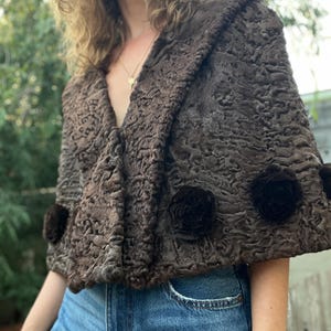 Pu&ograve; includere: Un mantello marrone strutturato con collo a scialle e tre rosette marrone scuro. Il mantello &egrave; realizzato in un materiale morbido, forse simile alla pelliccia. L'indumento &egrave; indossato sopra jeans blu.