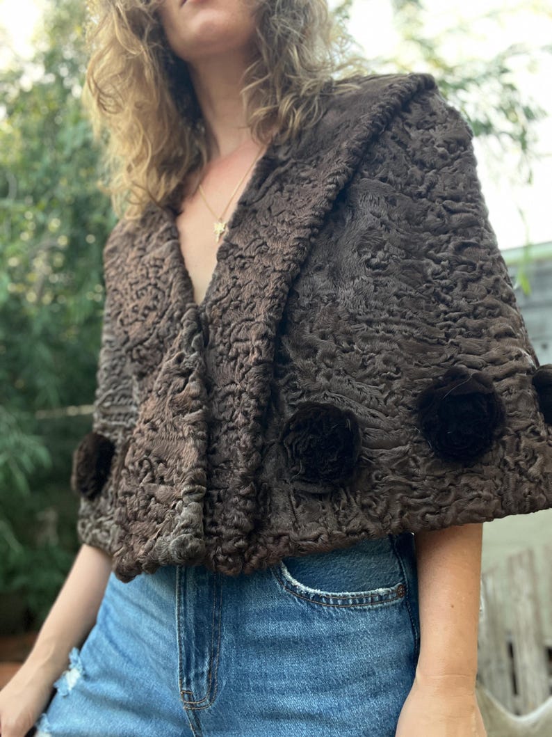 Pu&ograve; includere: Un mantello marrone strutturato con collo a scialle e tre rosette marrone scuro. Il mantello &egrave; realizzato in un materiale morbido, forse simile alla pelliccia. L'indumento &egrave; indossato sopra jeans blu.