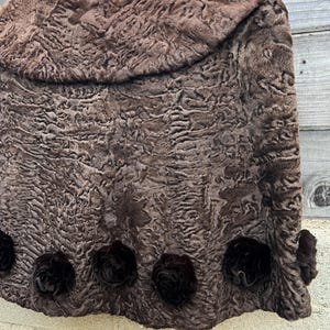 Pu&ograve; includere: Una mantella in pelliccia marrone, strutturata, con colletto arrotondato, appesa a una gruccia di legno. La mantella presenta rosette decorative marrone scuro lungo il bordo inferiore, aggiungendo un tocco di eleganza.