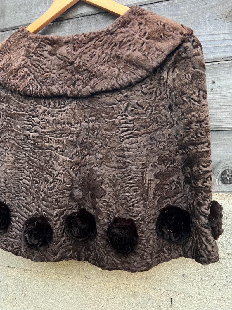 Pu&ograve; includere: Una mantella in pelliccia marrone, strutturata, con colletto arrotondato, appesa a una gruccia di legno. La mantella presenta rosette decorative marrone scuro lungo il bordo inferiore, aggiungendo un tocco di eleganza.
