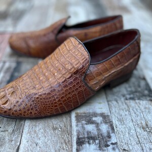 May include: A pair of brown leather slip-on shoes with a crocodile skin texture. The shoes have a rounded toe and a low heel. The background is a weathered wooden surface.