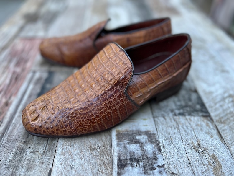 May include: A pair of brown leather slip-on shoes with a crocodile skin texture. The shoes have a rounded toe and a low heel. The background is a weathered wooden surface.