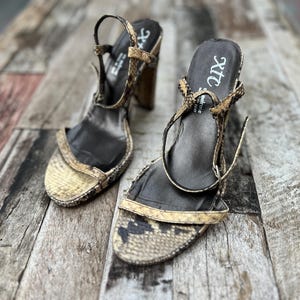 May include: A pair of high-heeled sandals with a snakeskin pattern in shades of tan, brown, and black. The sandals have a strap around the ankle and a block heel. The insole is black with the text "xtc".