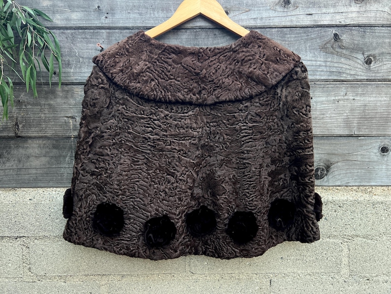 Pu&ograve; includere: Un mantello vintage marrone strutturato con colletto arrotondato e dettagli floreali marrone scuro. Il mantello &egrave; appeso a una gruccia di legno su uno sfondo di legno stagionato. Il tessuto del mantello ha una trama riccia unica.