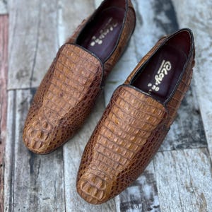 May include: A pair of brown leather loafers with a crocodile skin texture. The shoes have a classic slip-on design with a dark brown trim and a leather interior. The shoes are displayed on a wooden surface.