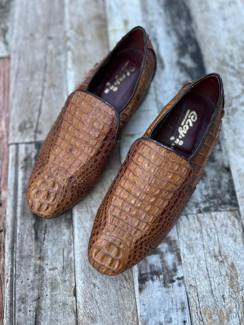 May include: A pair of brown leather loafers with a crocodile skin texture. The shoes have a classic slip-on design with a dark brown trim and a leather interior. The shoes are displayed on a wooden surface.