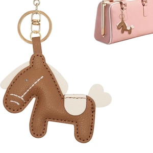 Peut inclure: Un porte-clés en forme de cheval marron avec un fermoir et un anneau en métal doré. Le cheval présente des accents blancs sur la crinière, la queue et la selle, avec des coutures blanches visibles sur les bords. Un petit sac rose est inclus.