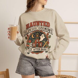 May include: A beige crewneck sweatshirt featuring a graphic print with the text "HAUNTED HOMEROOM SOCIETY EST. AFTER DARK". The design includes a schoolhouse, a ghost, books, an apple, a pencil, and autumn leaves in red, orange, and green.