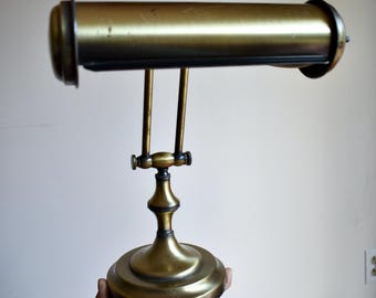 May include: A brass desk lamp featuring a cylindrical horizontal shade and a central adjustable arm connected to a circular base. The metal surface displays a polished finish with subtle signs of wear, including small dark spots and patina.