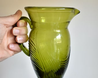 May include: A green glass pitcher with a handle and a spout. The pitcher has a textured, swirled design on the body. The pitcher is held by a hand.