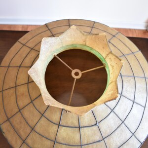 May include: Overhead view of a vintage lampshade. The shade has a circular design with a grid pattern and a star-shaped opening. The color is a mix of tan, beige, and brown, with a green accent around the opening. The metal frame is visible.