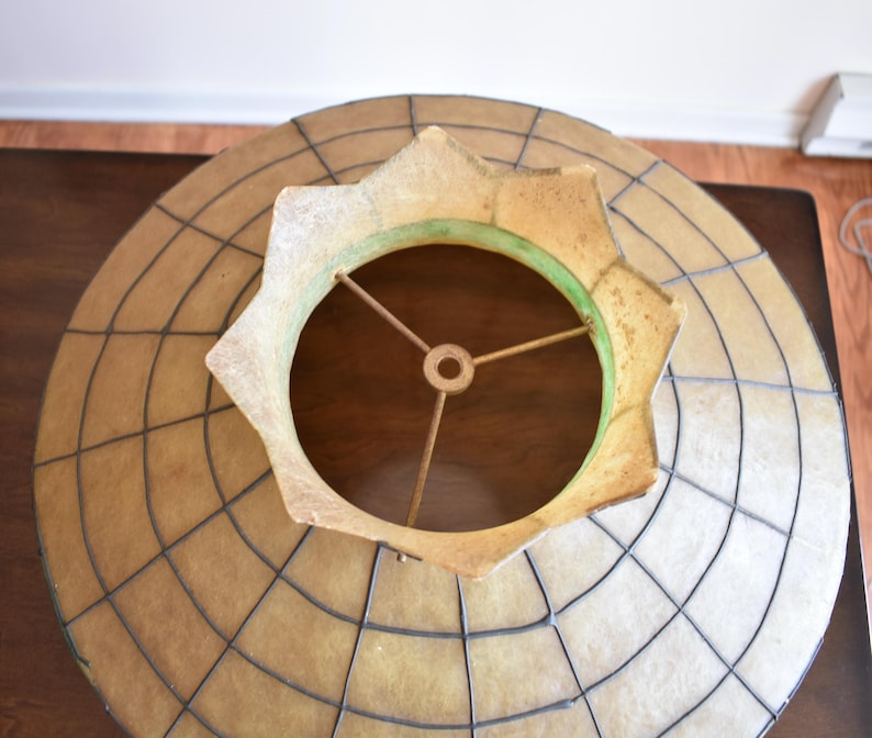 May include: Overhead view of a vintage lampshade. The shade has a circular design with a grid pattern and a star-shaped opening. The color is a mix of tan, beige, and brown, with a green accent around the opening. The metal frame is visible.