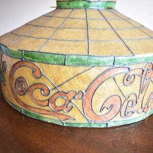 May include: A vintage Coca-Cola stained glass lamp shade. The shade features the Coca-Cola logo in orange and brown against a yellow background. The design is framed with green and black lines.