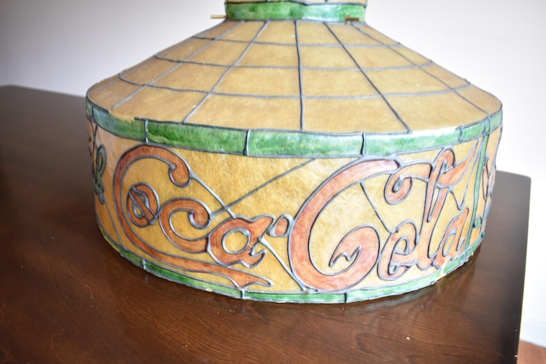 May include: A vintage Coca-Cola stained glass lamp shade. The shade features the Coca-Cola logo in orange and brown against a yellow background. The design is framed with green and black lines.