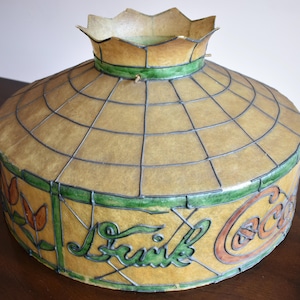 May include: Vintage Coca-Cola lamp shade with a yellow and green stained glass design. The shade features the Coca-Cola logo and the word "Drink" in green script. The shade has a grid pattern and a scalloped top.