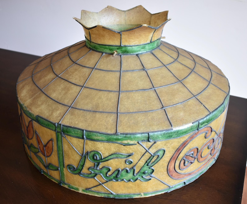 May include: Vintage Coca-Cola lamp shade with a yellow and green stained glass design. The shade features the Coca-Cola logo and the word "Drink" in green script. The shade has a grid pattern and a scalloped top.