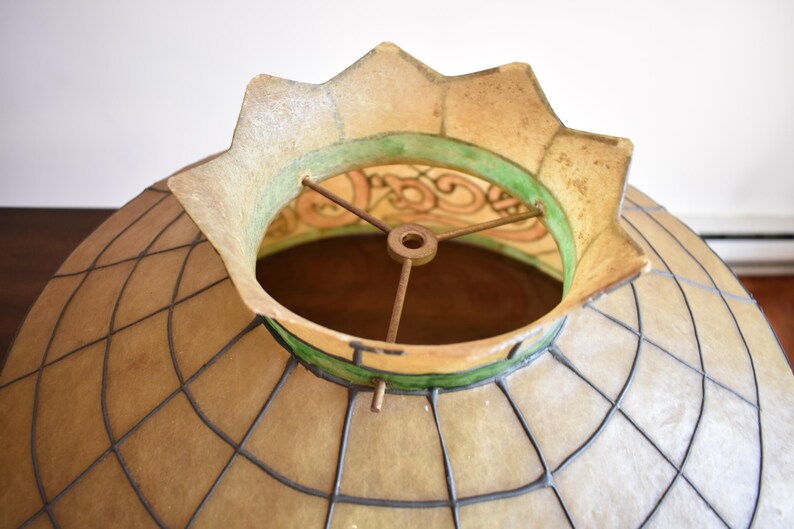 May include: A dome-shaped lamp shade featuring a grid of dark metal lines over translucent beige panels. The top opening has a scalloped edge with a green band and a metal spider fitter with three spokes centered around a central ring.