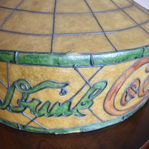 May include: Vintage Coca-Cola stained glass lamp shade. The shade features a globe-like shape with a grid pattern. The Coca-Cola logo is displayed in green and orange lettering. The shade is trimmed with green glass.