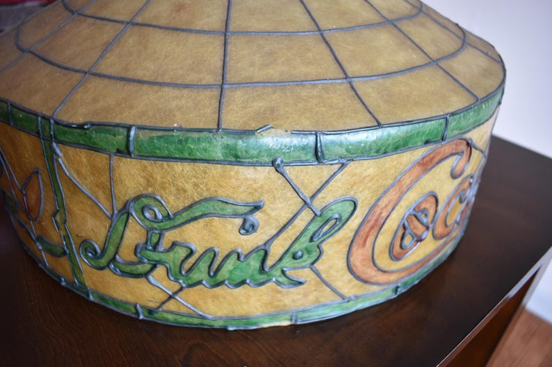 May include: Vintage Coca-Cola stained glass lamp shade. The shade features a globe-like shape with a grid pattern. The Coca-Cola logo is displayed in green and orange lettering. The shade is trimmed with green glass.