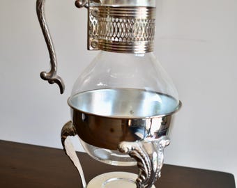 May include: A clear glass carafe with a silver-toned metal frame, featuring an ornate handle, a decorative filigree collar, and a lid. The carafe rests on a three-legged silver-toned metal stand with a circular base and detailed scrollwork.