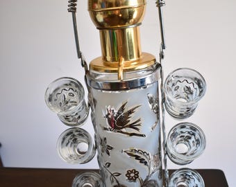 May include: Vintage cocktail shaker with six shot glasses. The shaker has a gold-colored top and a silver-colored body with etched bird and floral designs. A silver handle arches over the top.