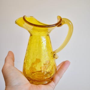 Puede incluir: Una jarra de vidrio amarilla con un acabado craquelado. La jarra tiene un asa y un pico acampanado. La jarra se sostiene en una mano sobre un fondo liso. La jarra probablemente se utiliza para servir bebidas.