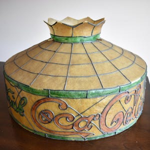 May include: A vintage Coca-Cola stained glass lamp shade. The shade features a domed shape with a grid pattern and the Coca-Cola logo in brown and green. The top has a scalloped edge.