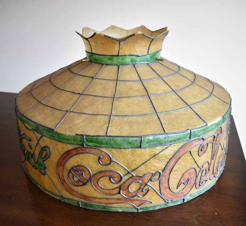 May include: A vintage Coca-Cola stained glass lamp shade. The shade features a domed shape with a grid pattern and the Coca-Cola logo in brown and green. The top has a scalloped edge.