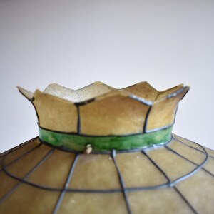 May include: Close-up of a vintage lampshade with a scalloped edge. The shade is a light tan color with a green band and a grid pattern. The lampshade is likely made of glass or a similar material.