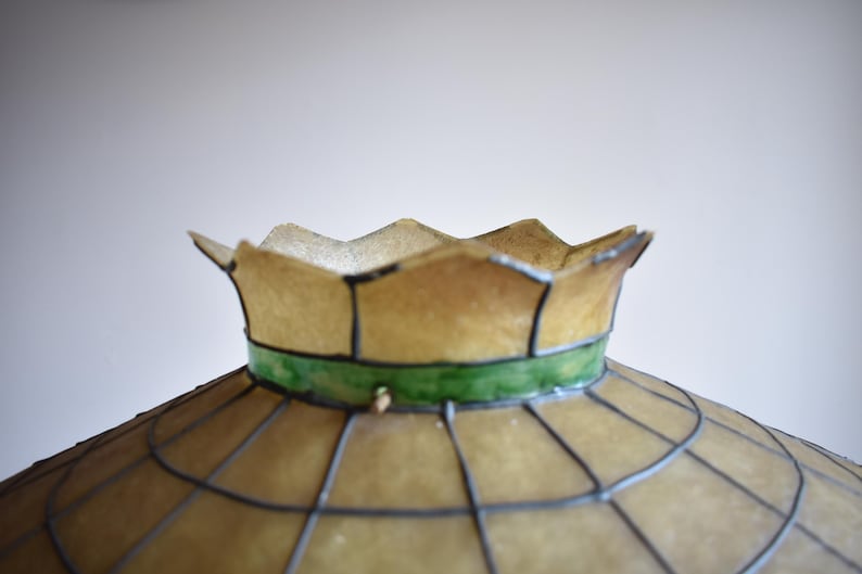May include: Close-up of a vintage lampshade with a scalloped edge. The shade is a light tan color with a green band and a grid pattern. The lampshade is likely made of glass or a similar material.