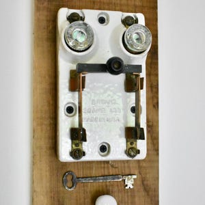 May include: Vintage electrical switch mounted on a wooden board. The white switch has two glass-covered components and copper contacts. A small key and a white ceramic knob are also attached to the wood. The switch is marked "BRYANT 20 AMP 125V MADE IN USA."
