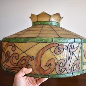 May include: Vintage Coca-Cola stained glass lamp shade. The shade features the Coca-Cola logo in brown and black, with green and tan accents. The shade has a crown-like top.