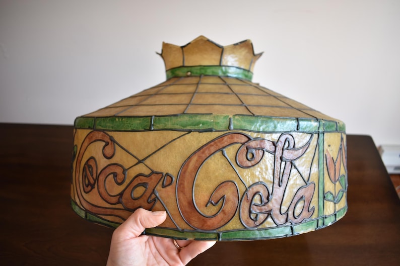 May include: Vintage Coca-Cola stained glass lamp shade. The shade features the Coca-Cola logo in brown and black, with green and tan accents. The shade has a crown-like top.
