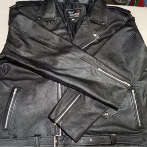 Könnte beinhalten: Eine schwarze Lederjacke mit Reverskragen, silberfarbenen Metallreißverschlüssen an der Vorderseite und an den Ärmelbündchen sowie einem passenden Ledergürtel am Saum. Das Innenetikett zeigt Real Leather Made in Pakistan. Eine Metallschere liegt daneben.