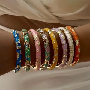 Pode incluir: Uma coleção de oito pulseiras rígidas articuladas em várias cores, incluindo azul, verde, rosa, multicolorido, amarelo, lavanda, branco e laranja. Cada pulseira de metal apresenta um padrão repetitivo de flores e folhas douradas.