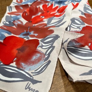 May include: A silk scarf with a floral design. The scarf displays large red flowers, grey leaves, and light blue accents on a cream background. The scarf has the signature "Usna" in the corner.