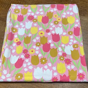May include: A folded pink fabric featuring a repeating floral pattern of tulips and small flowers. The tulips are in white, yellow, pink, and brown, with green stems and leaves. The small flowers are yellow and pink.