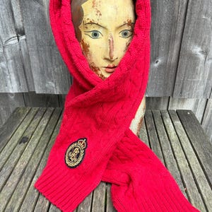 May include: A bright red cable-knit hooded scarf featuring a ribbed hem and a circular embroidered patch on the front. The patch displays a gold-coloured crest with a crown and intertwined letters, set against a dark background with a gold border.