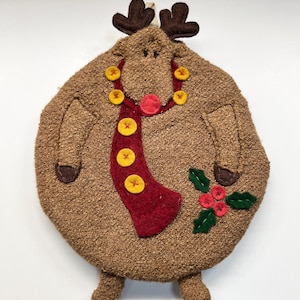May include: A round, textured brown fabric ornament shaped like a reindeer. It features dark brown felt antlers, a red felt scarf adorned with five yellow buttons, a red nose, and a green holly leaf applique with three red berries on the side.