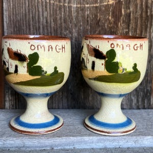 May include: Two cream-coloured ceramic egg cups featuring a hand-painted landscape scene with green hills, brown structures, and the text OMAGH in brown lettering. Each cup has a brown rim and a blue band around the base with a visible crackle glaze finish.