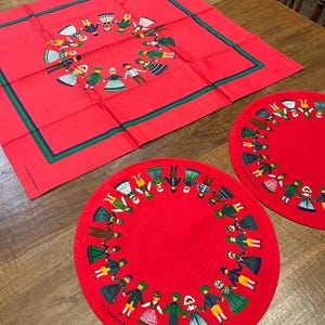 May include: A red tablecloth and two matching placemats with a green border. Each item has a circular design of children holding hands. The children are dressed in various colours, creating a festive and playful aesthetic.